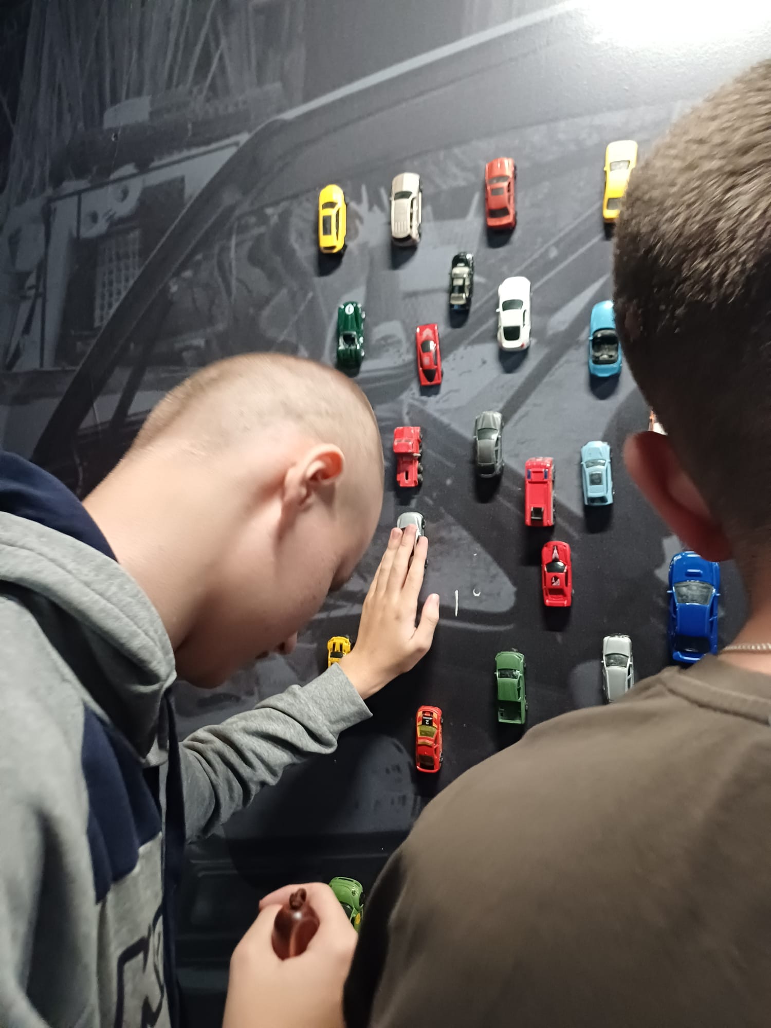 Dva učenika stoje okrenuta zidu s nizovima malih šarenih autića; jedan učenik u sivoj hoodici dodiruje autić rukom, drugi je djelomično u kadru.